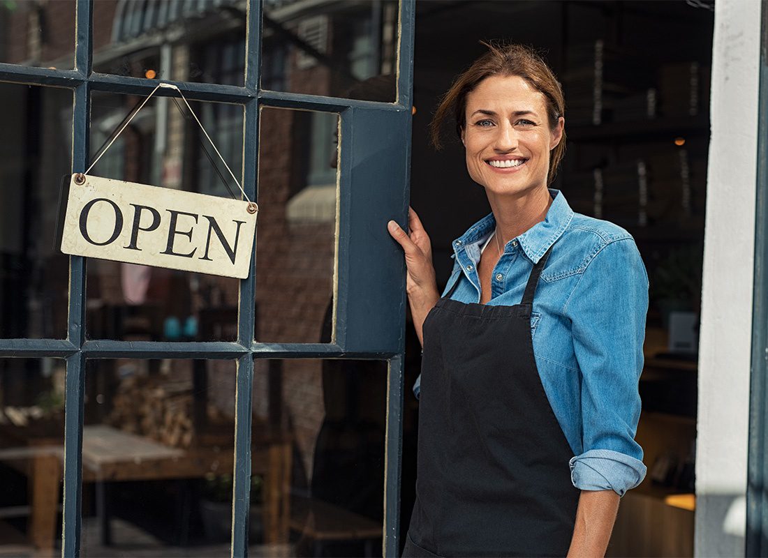Business Insurance - Portrait of a Smiling Middle Aged Business Woman Standing Next to the Door of her Main Street Shop with an Open Sign Hanging on the Front
