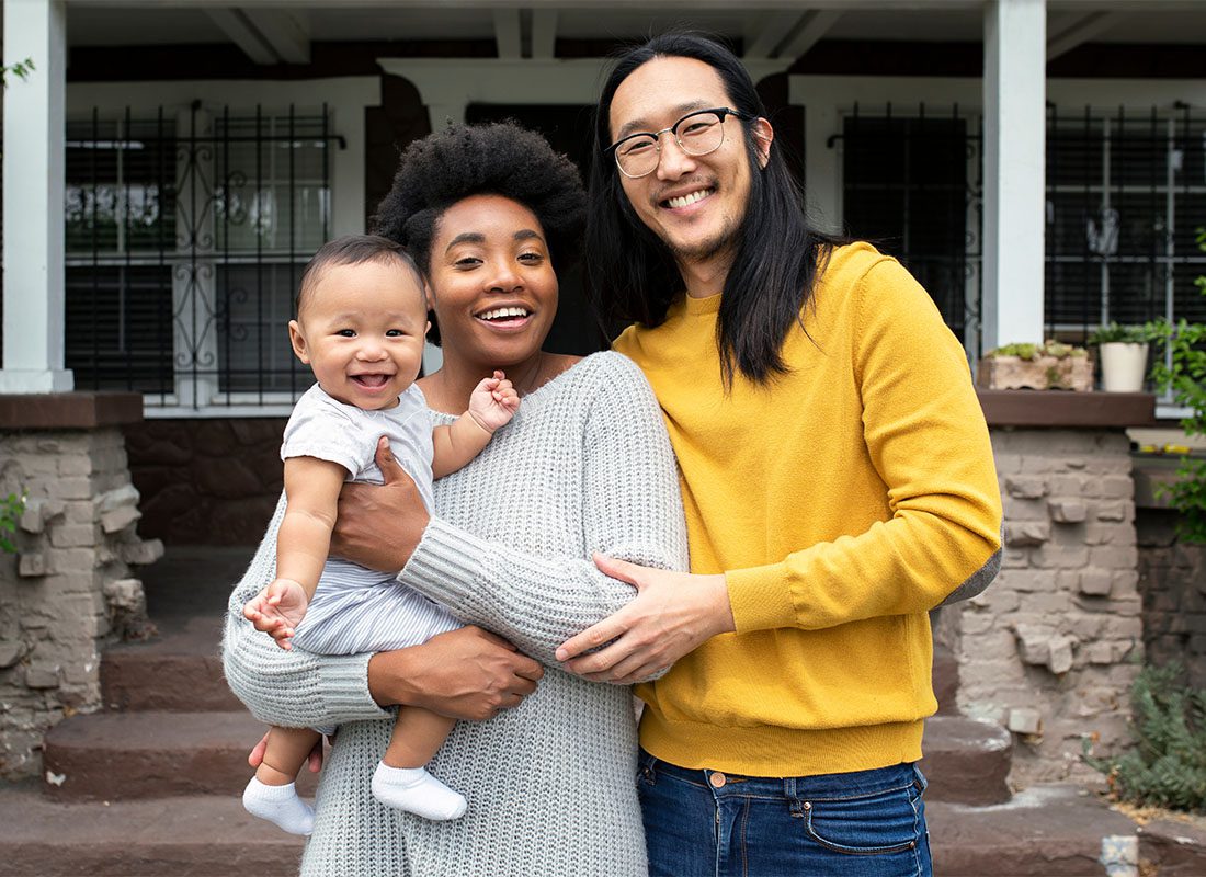 Personal Insurance - Portrait of a Cheerful Young Family Holding Their Baby as They Stand Outside Their New Home